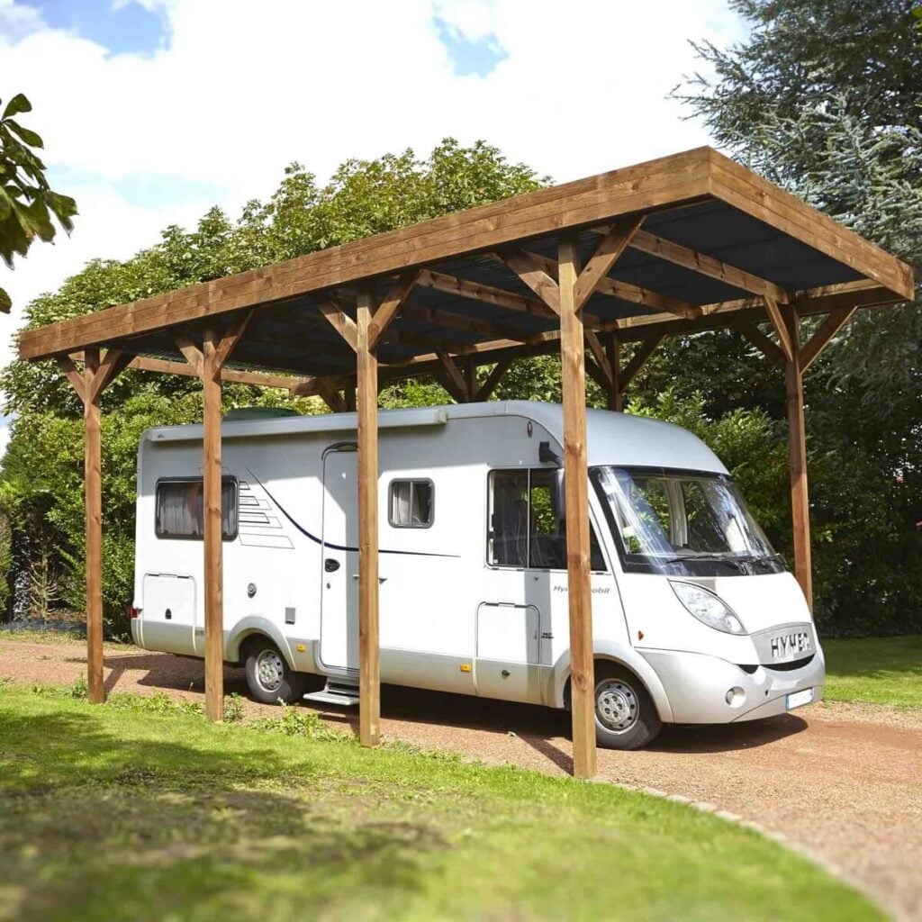 Carport en bois avec camping-car garé, sur allée gravillonnée entourée d'arbres